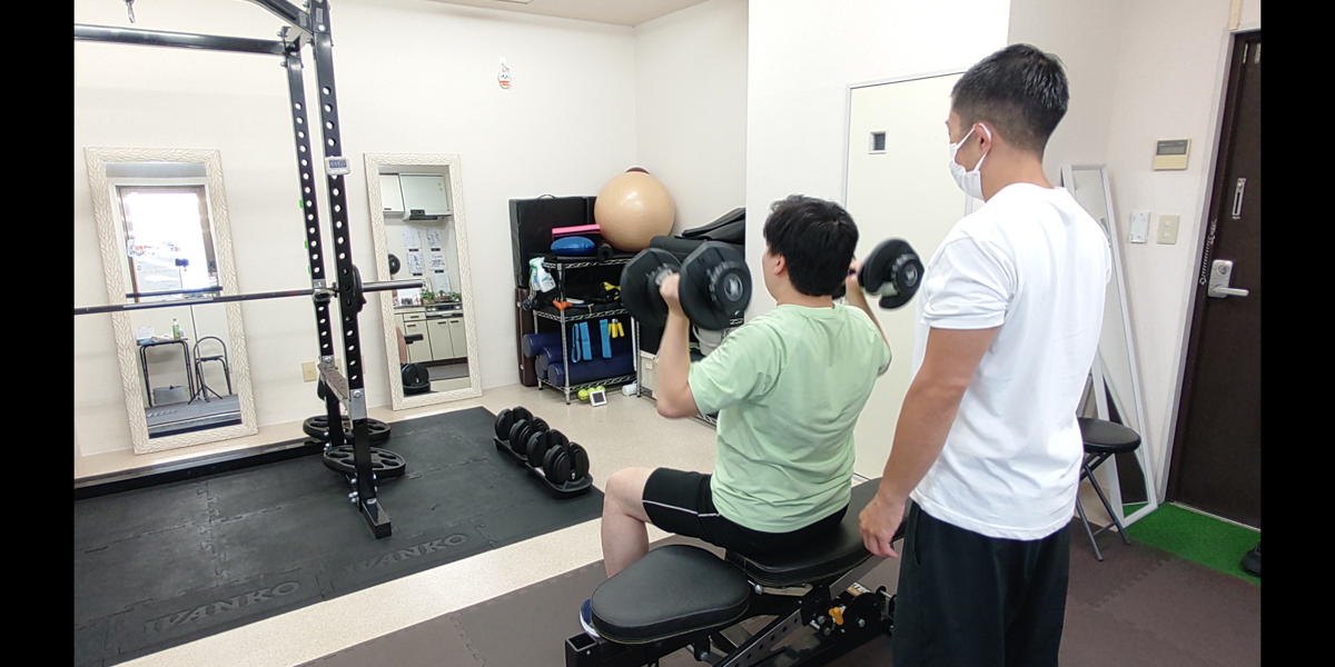 トレーナーが見守る中でダンベルショルダープレスを行うお客様