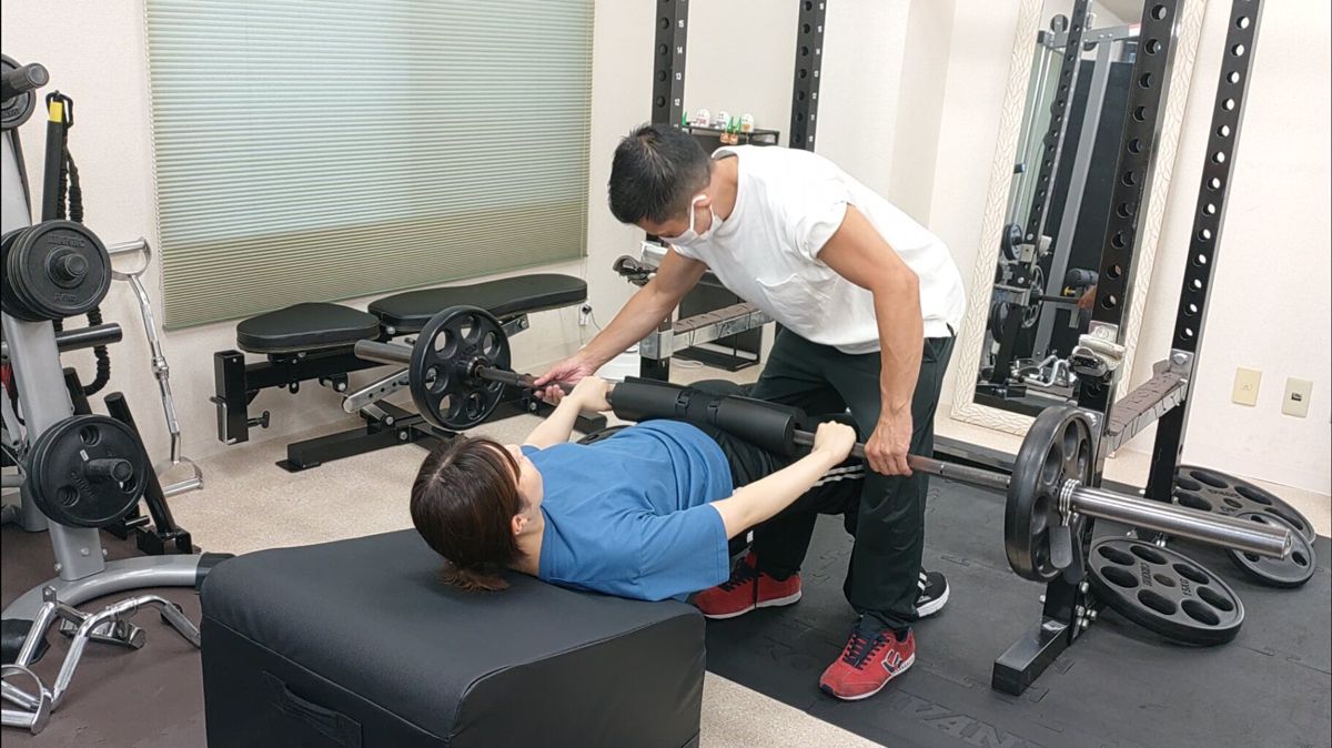 トレーナーがバーベルを支えながらヒップスラストをサポートする様子